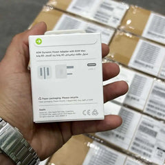 Hand holding a white power adapter box with 'Apple' branding, stacked boxes in the background.

